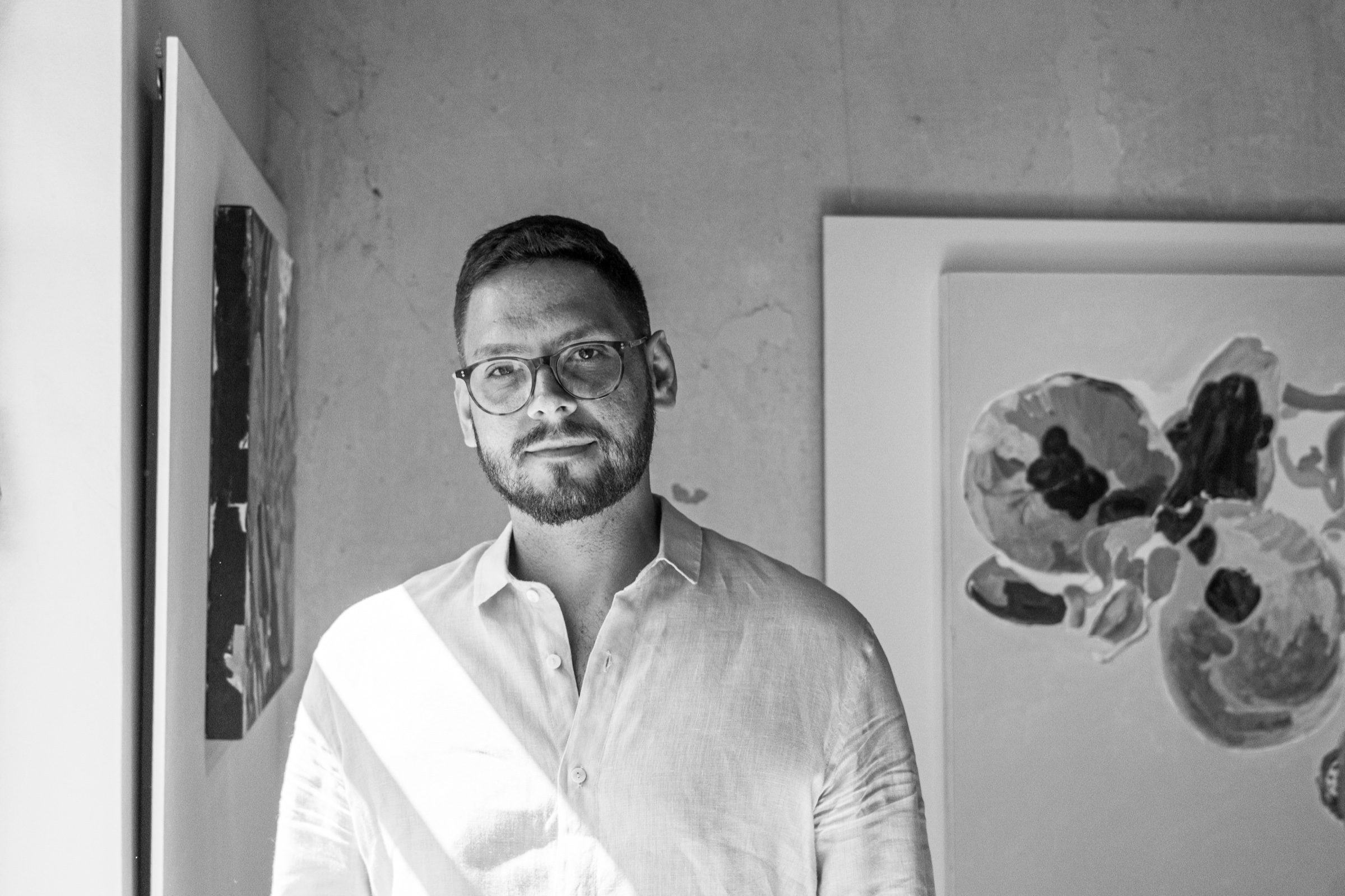 Retrato em preto e branco de Gustavo Faria diante de obras de arte.